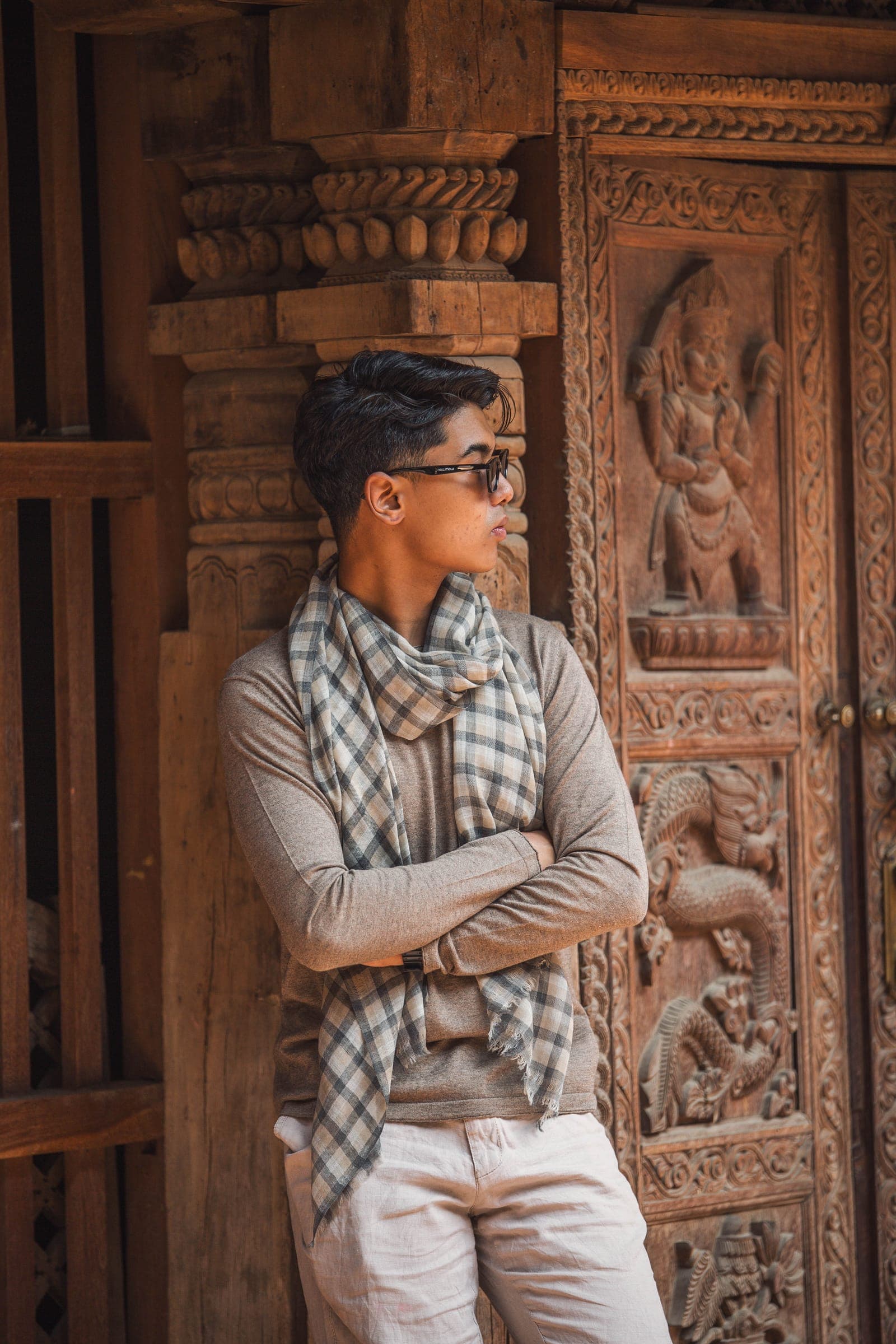 A man in a grey sweater and checked cashmere scarf leans against a carved wooden temple door