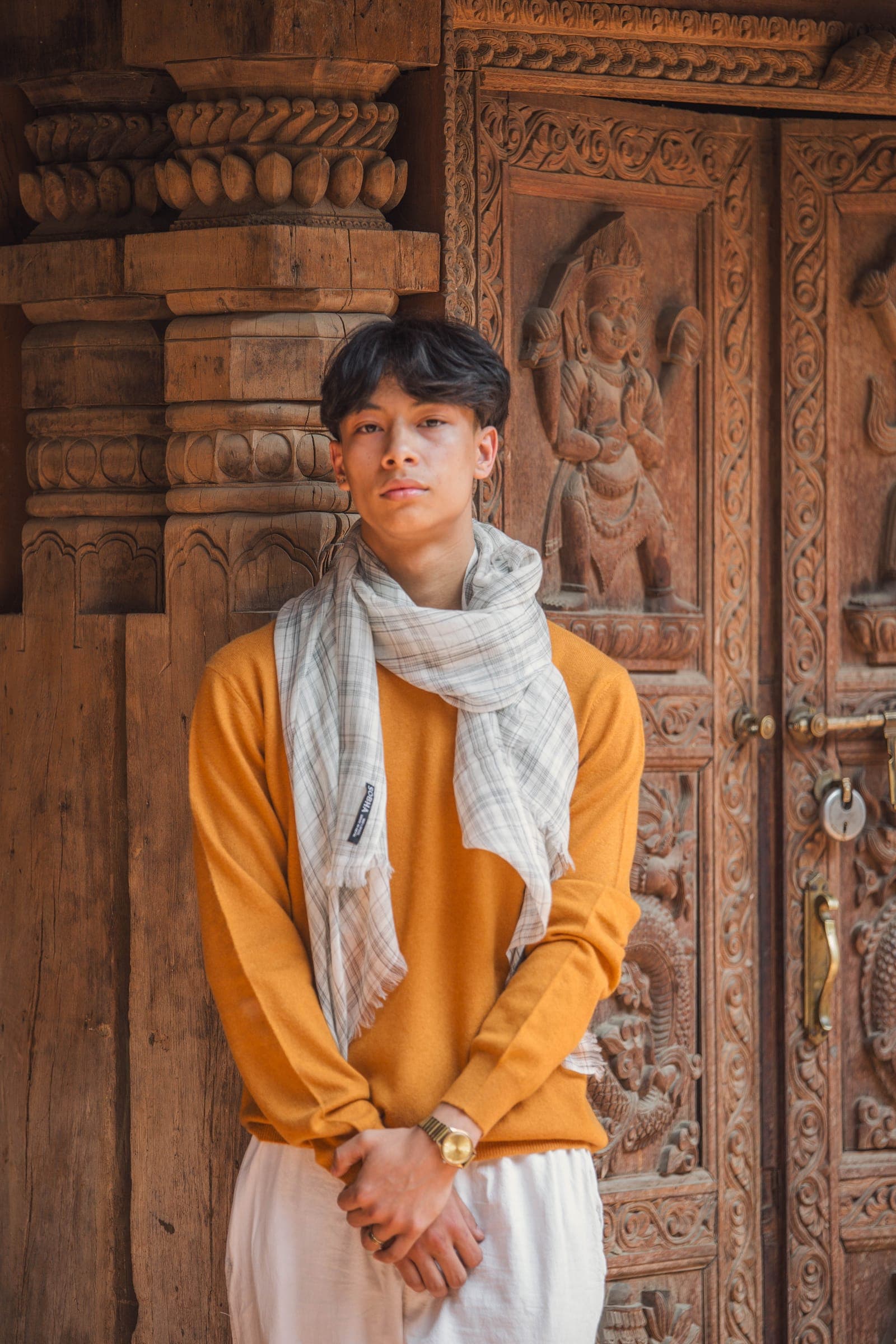 A young man in a mustard sweater and pale checked scarf stands before a carved wooden door with brass handles