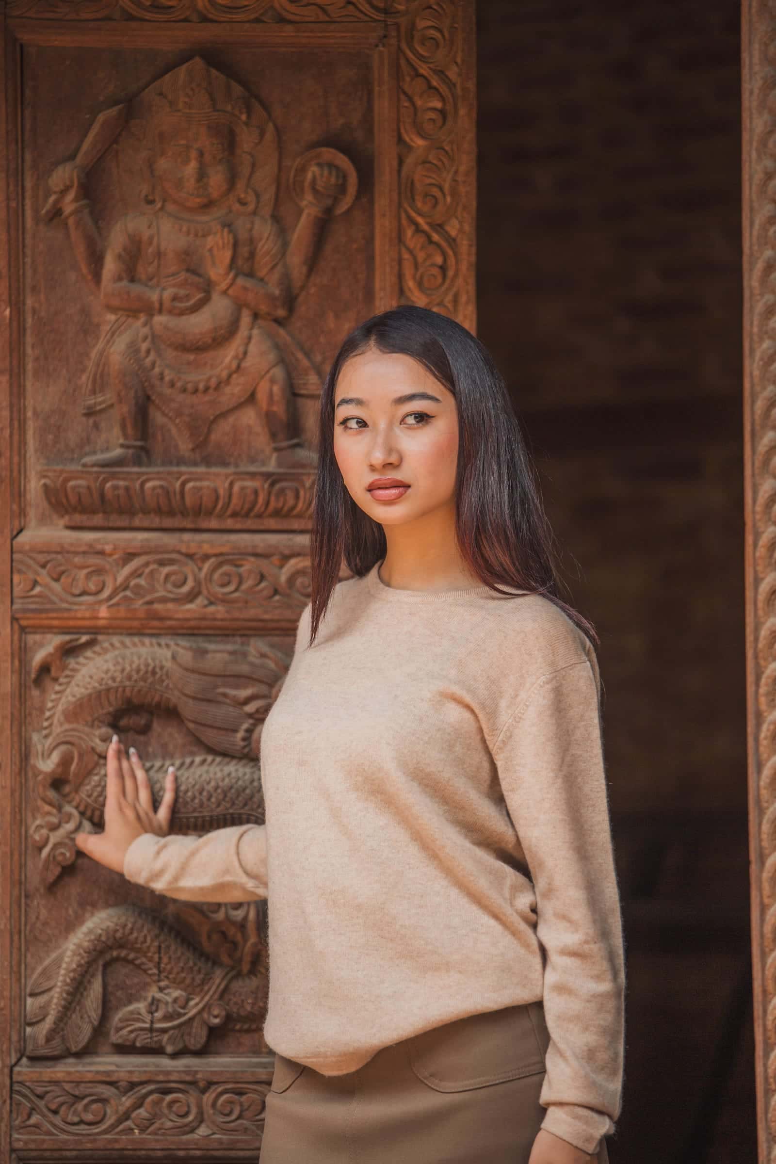 A woman in a beige cashmere sweater rests her hand on a wooden panel carved with deities and serpents
