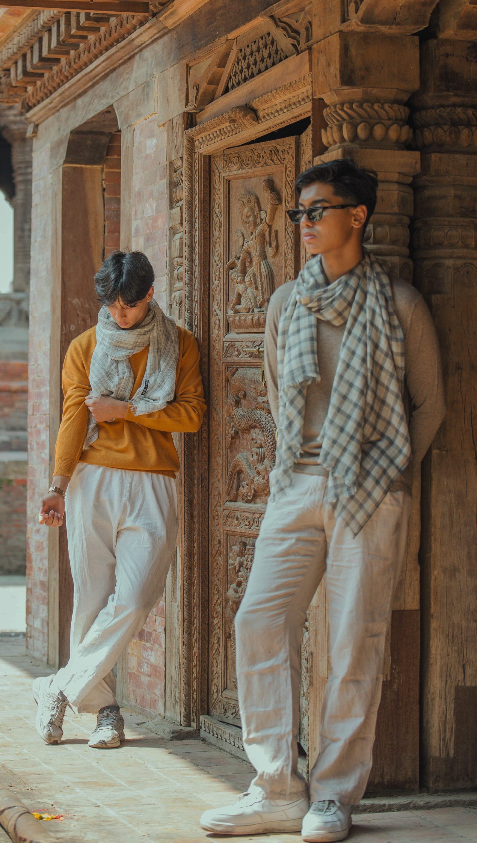 Two young men in checked cashmere scarves lean against carved wooden doors in a brick courtyard