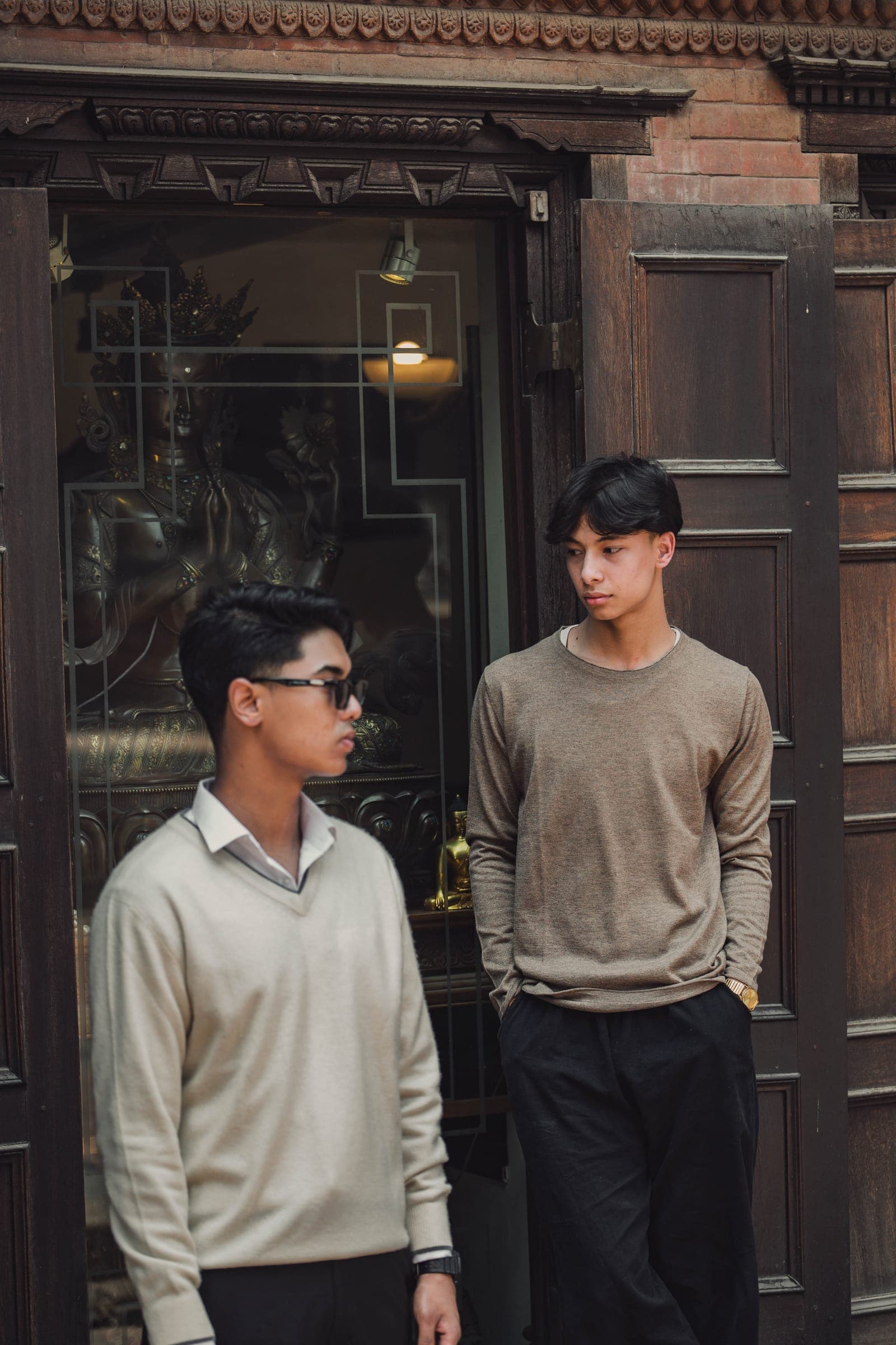 Two men in cream and taupe cashmere sweaters talk in a doorway framed by carved wood and brick