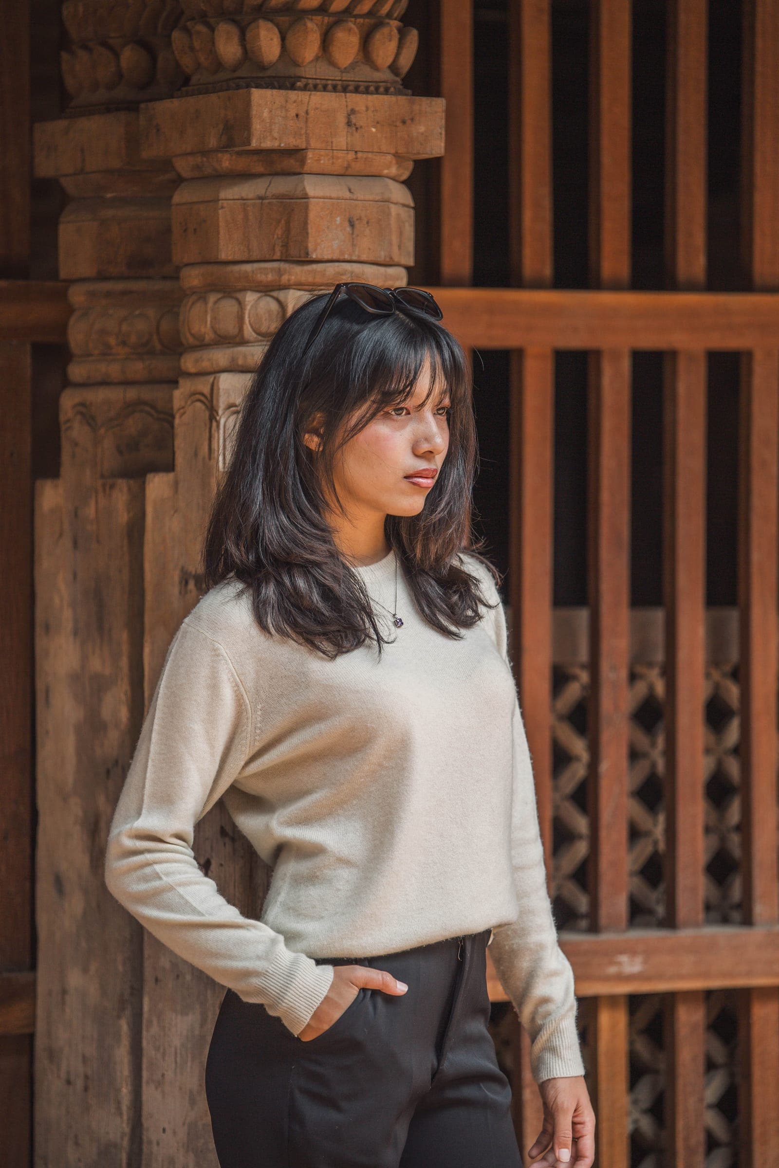 A woman in a cream cashmere sweater stands beside a carved pillar and a wooden lattice window