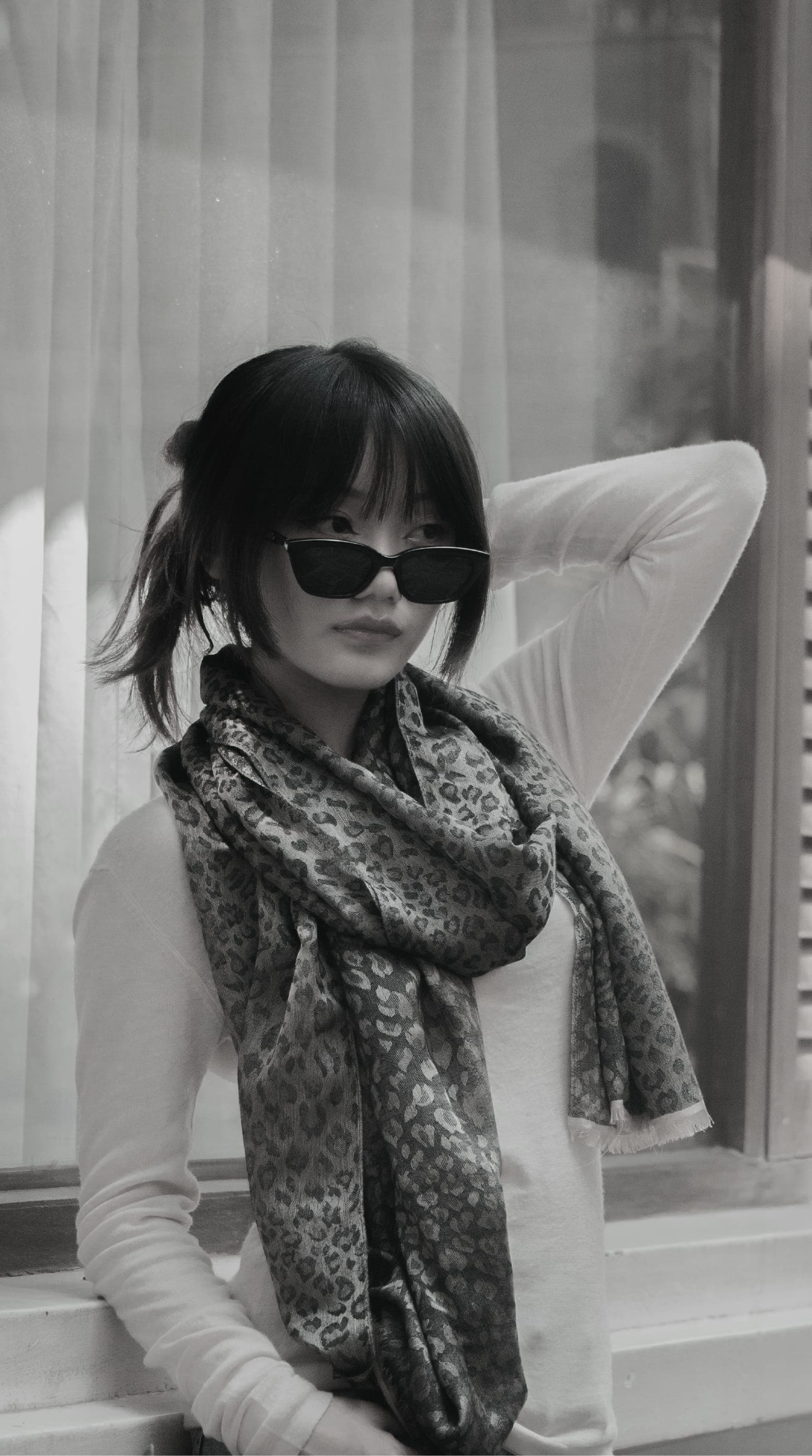 Black-and-white portrait of a woman in sunglasses wearing a leopard-pattern cashmere scarf by a window