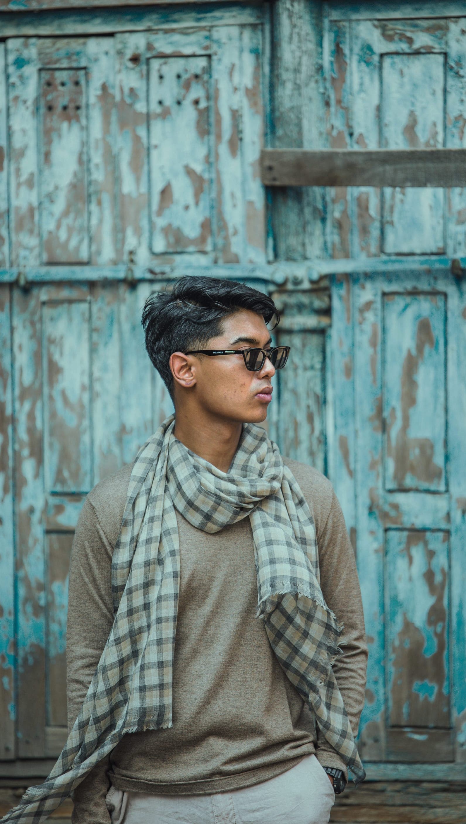 A man in a taupe sweater and checked cashmere scarf in front of a weathered turquoise wooden door